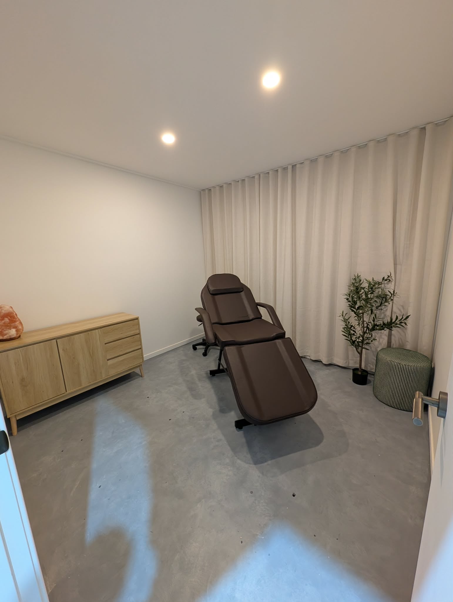 A brown massage chair positioned in a room with a concrete floor, beige curtains, a light wood cabinet, and a small potted plant.