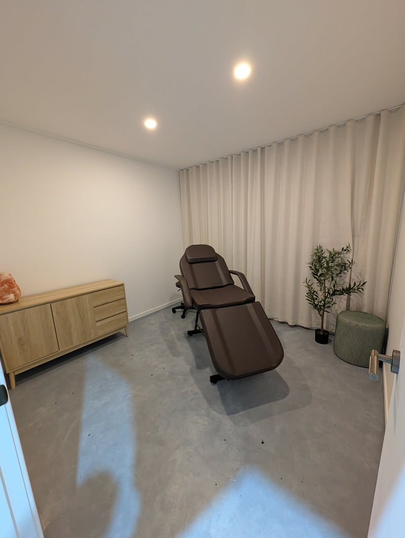 A brown massage chair positioned in a room with a concrete floor, beige curtains, a light wood cabinet, and a small potted plant.