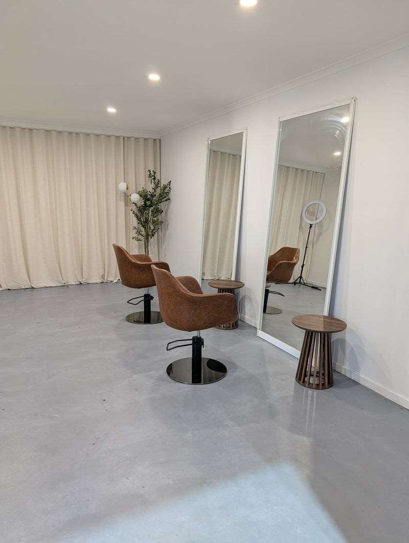 A modern salon interior with two brown velvet swivel chairs, two full-length mirrors, a small round wooden stool, and cream curtains in the background.