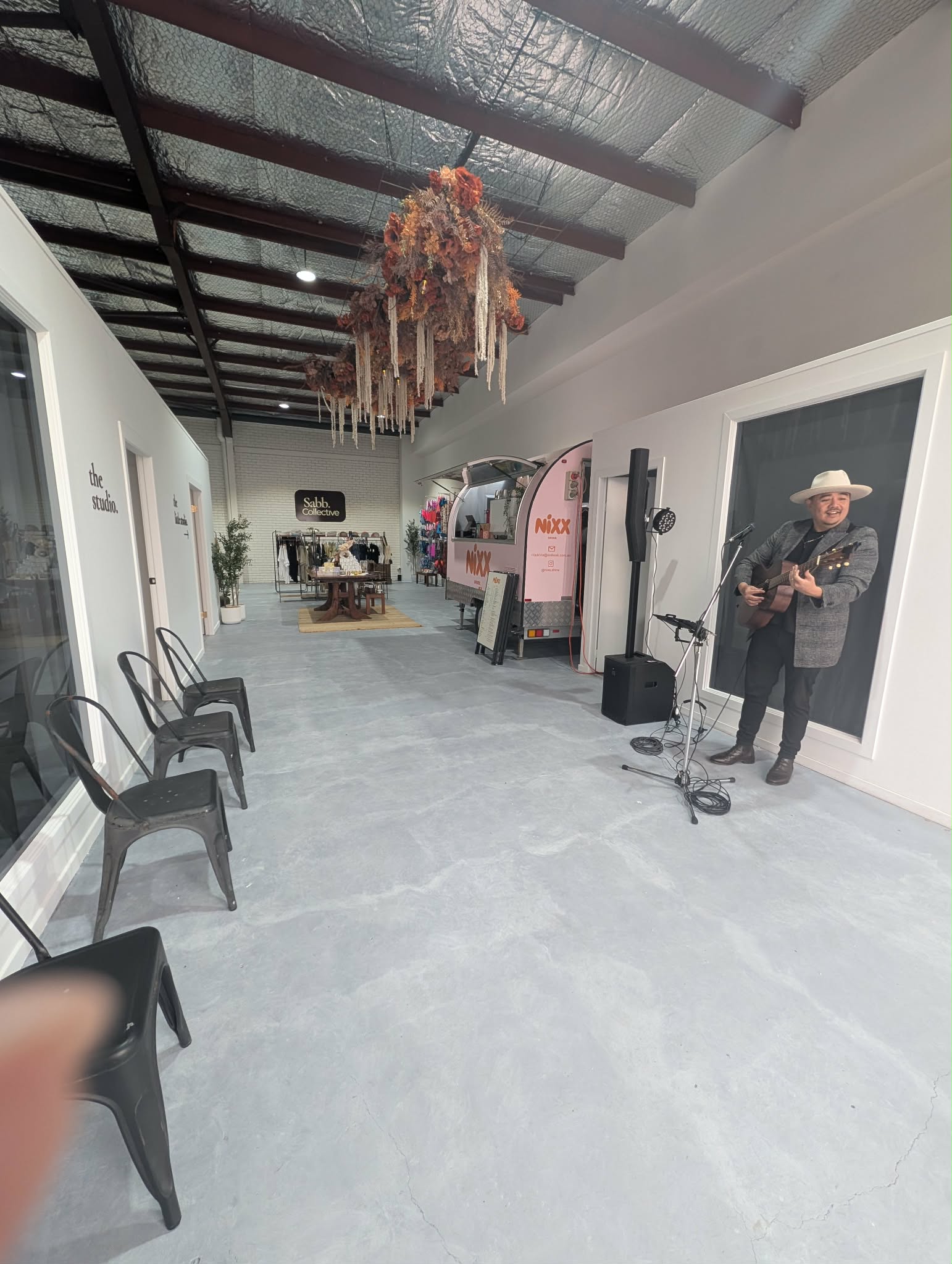 A person wearing a cowboy hat and dark clothing stands near a microphone and speakers in a long, open indoor space with a concrete floor, black chairs, exposed ceiling beams, and a decorative hanging arrangement overhead.