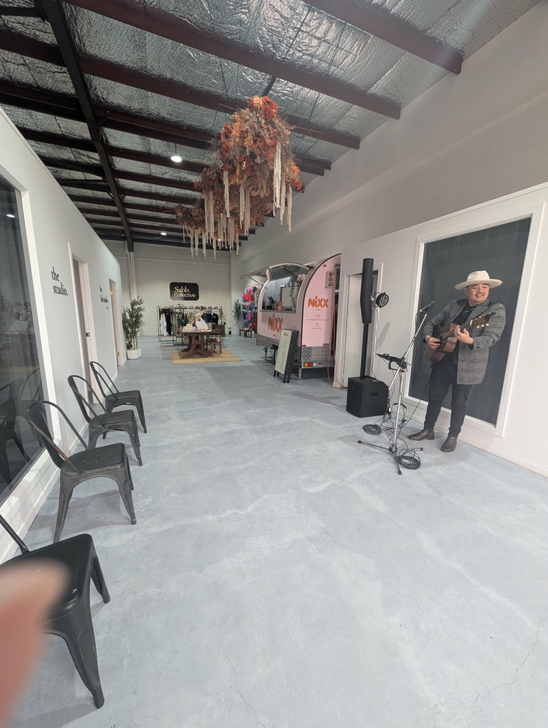 A person wearing a cowboy hat and dark clothing stands near a microphone and speakers in a long, open indoor space with a concrete floor, black chairs, exposed ceiling beams, and a decorative hanging arrangement overhead.