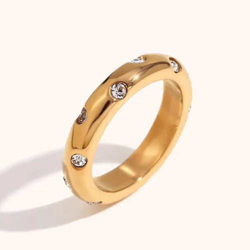 A polished yellow gold band ring featuring small, evenly spaced bezel-set clear round gemstones, photographed on a clean off-white background with a soft shadow.