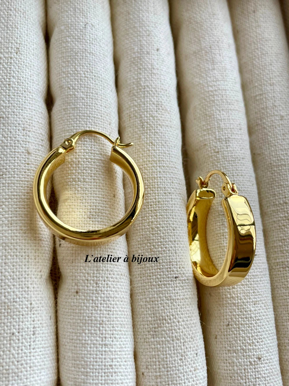 A pair of polished gold hoop earrings displayed on a textured beige fabric jewelry stand. One earring is shown from the front, and the other is angled to show its side profile and latch mechanism.
