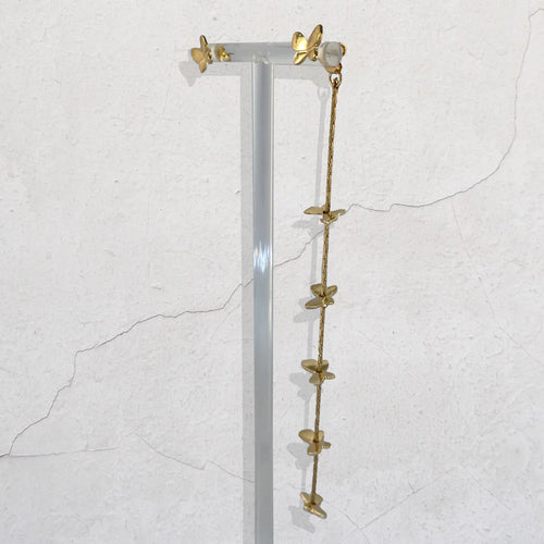 A long gold-toned dangle necklace with multiple small butterfly-shaped charms hangs from a clear acrylic display stand against a textured off-white wall.