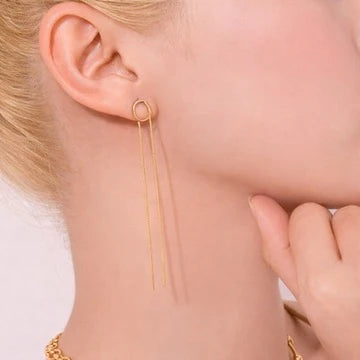 A close-up of a model's ear wearing a delicate gold dangle earring with a small circular hoop and two long thin drops, against a soft light background.
