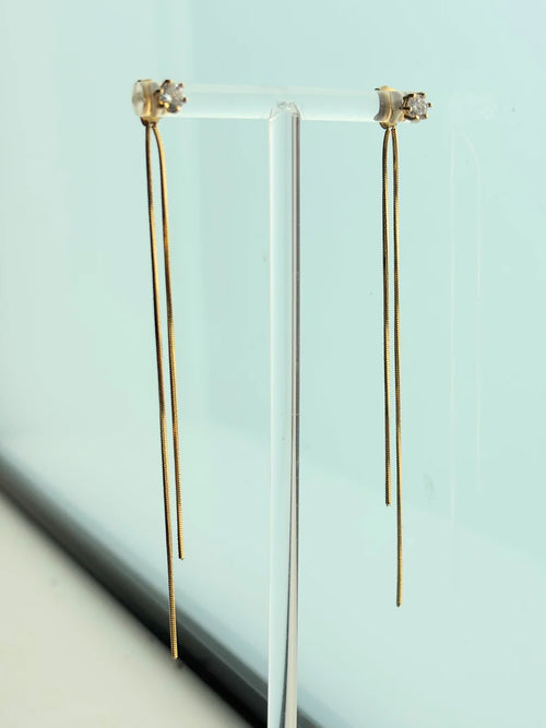 A pair of long, delicate gold dangle earrings displayed on a clear acrylic stand against a pale blue background.