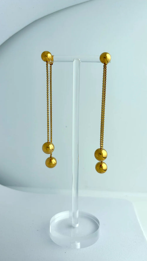 A pair of gold dangling earrings with spherical bead details, displayed on a clear acrylic stand against a light gray background.