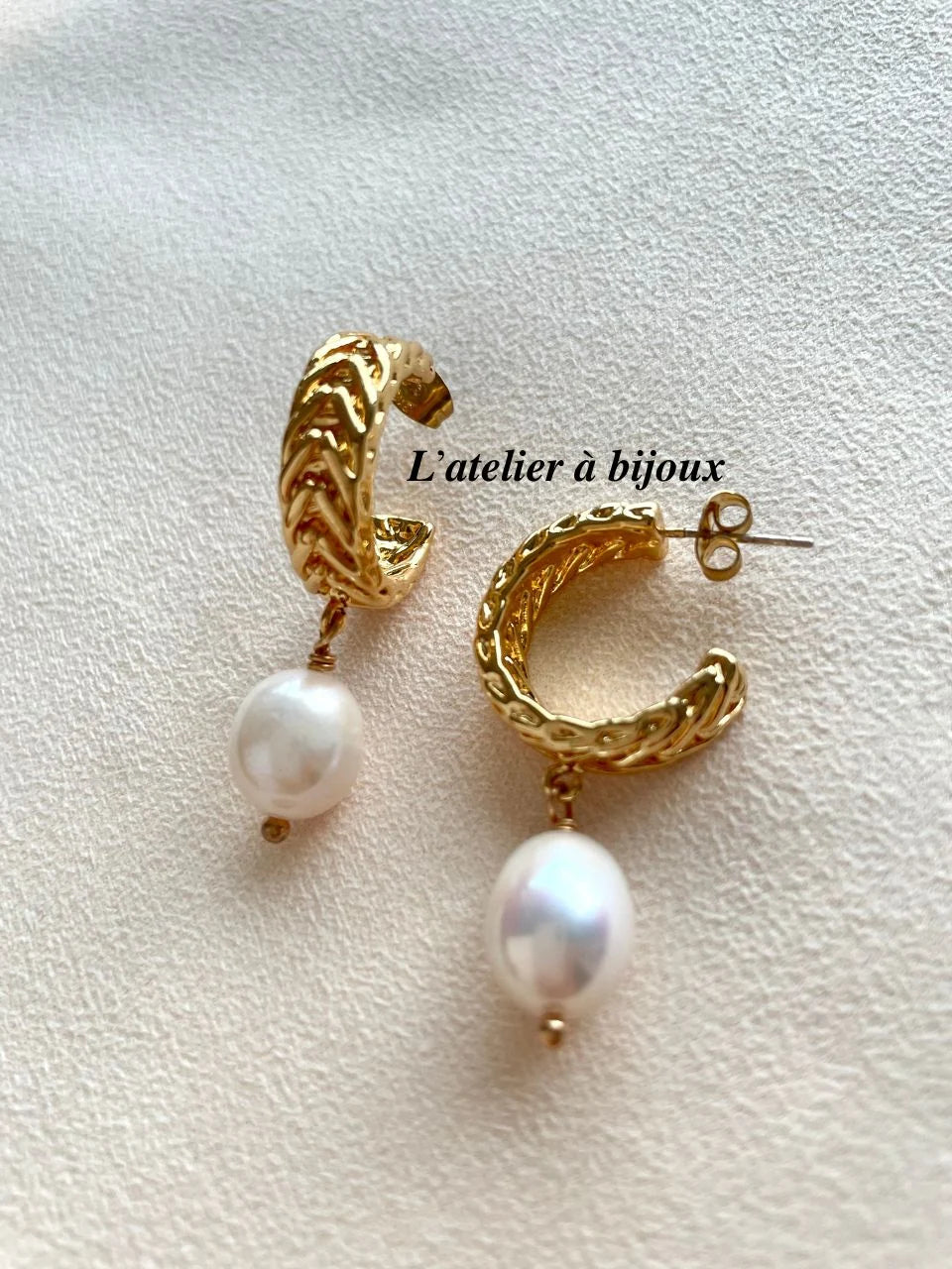 A pair of gold huggie hoop earrings with a textured chevron pattern and a dangling white pearl, displayed on a textured cream fabric background with a watermark 'L'atelier à bijoux'.