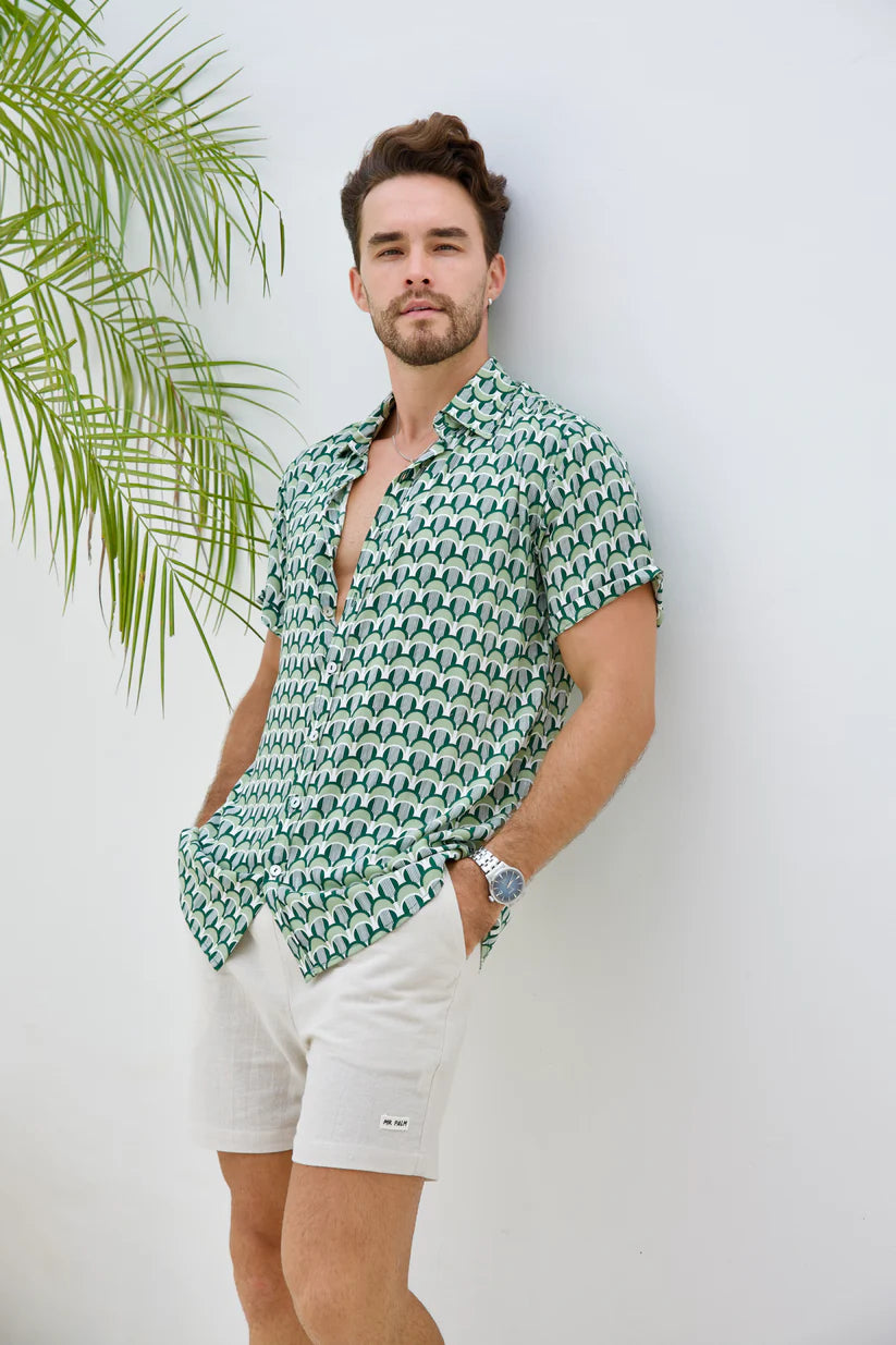 A man wearing a green and white patterned short-sleeve button-up shirt and white shorts, leaning against a white wall with a palm leaf on the left.