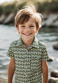 A young boy smiling outdoors near the water, wearing a short-sleeve button-up shirt with a repeating green, beige, and white arch pattern.