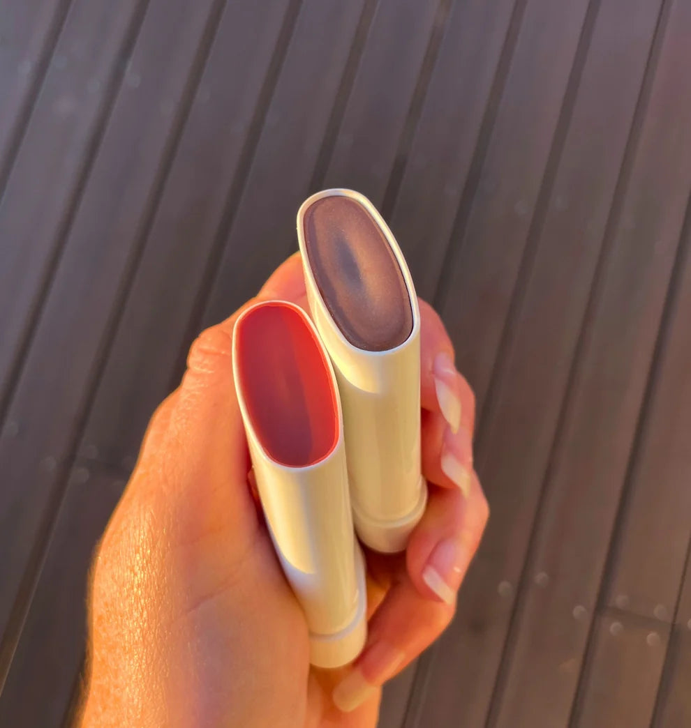A hand holds two cream-colored lip balm tubes outdoors against a dark wooden slatted background. One balm stick is open, showing a pinkish-red balm inside, and the other is closed, showing a mauve-brown balm surface.