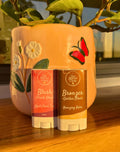 Two cosmetic sticks labeled 'Blush' and 'Bronzer' in front of a decorative vase with floral design.