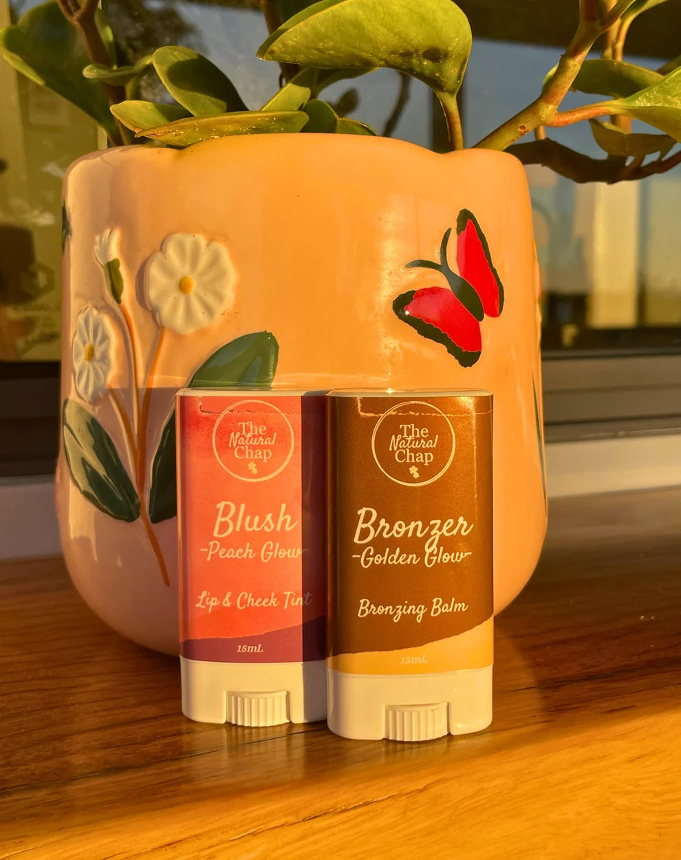 Two cosmetic sticks labeled 'Blush' and 'Bronzer' in front of a decorative vase with floral design.