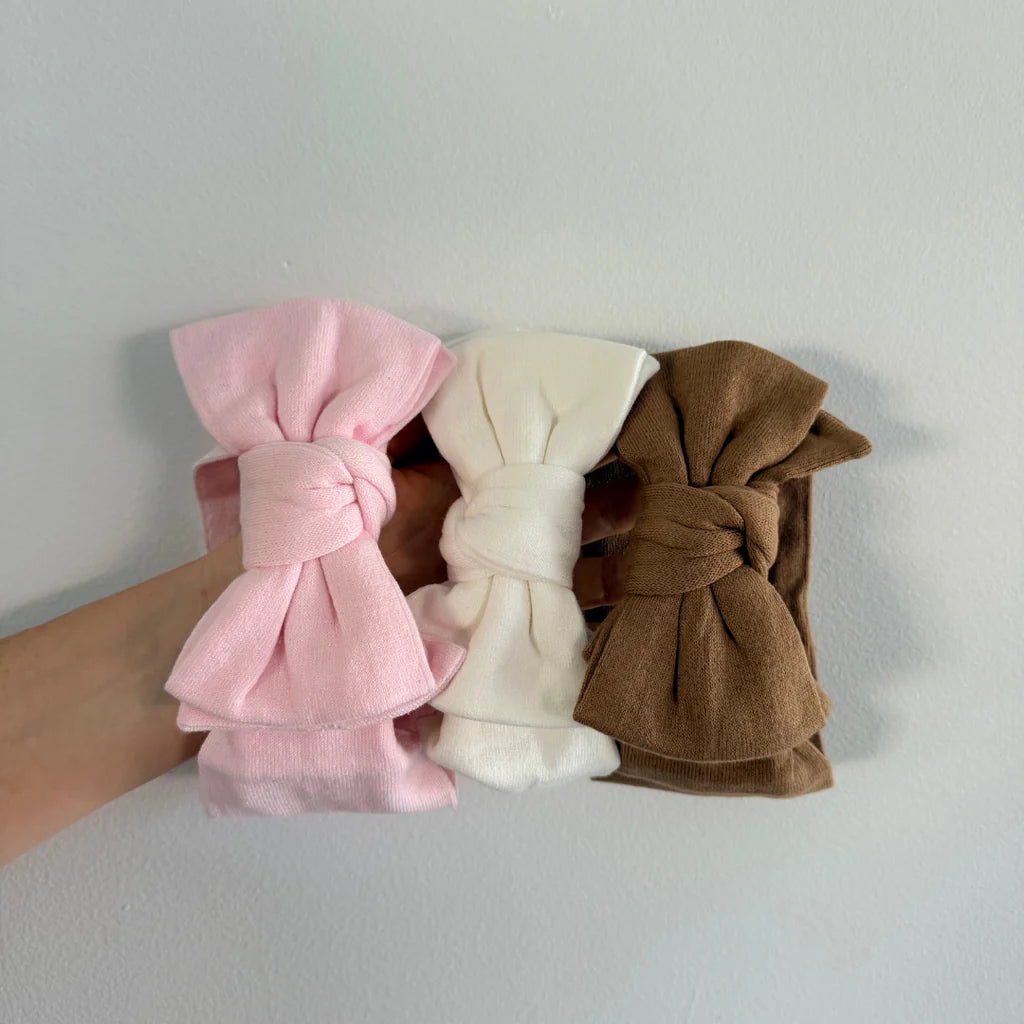 Three fabric bow hair clips in pink, cream, and brown, held in a hand against a plain light gray wall.