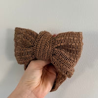 A hand holding a large, textured brown crocheted bow headband against a plain off-white wall.