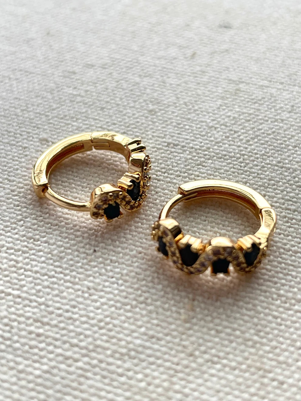A pair of small gold huggie hoop earrings with black enamel detailing, displayed on a textured white fabric background.