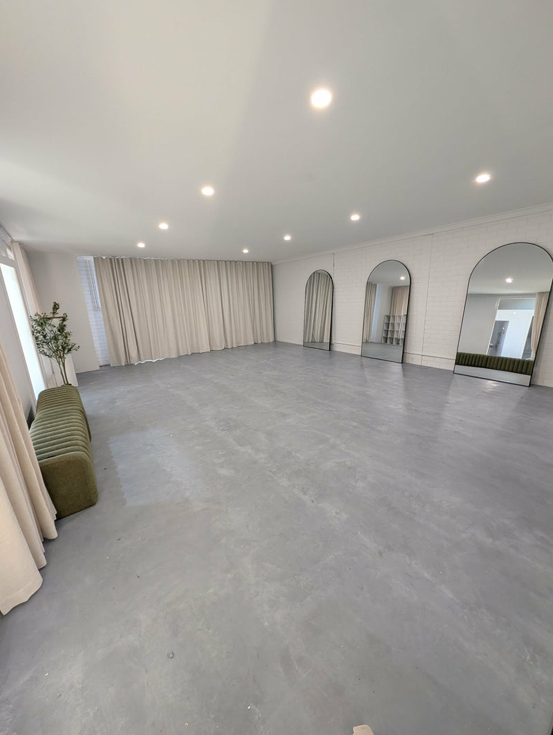 A large, empty studio room with a gray concrete floor, white walls, recessed ceiling lights, cream curtains, and arched mirrors. A small green bench and potted plant are visible on the left side.