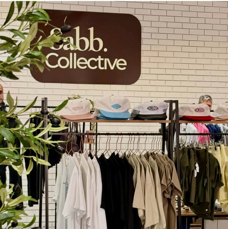 A retail display for Sabb Collective featuring a large brown brand sign on a white brick wall, with racks of t-shirts and several baseball caps displayed below.