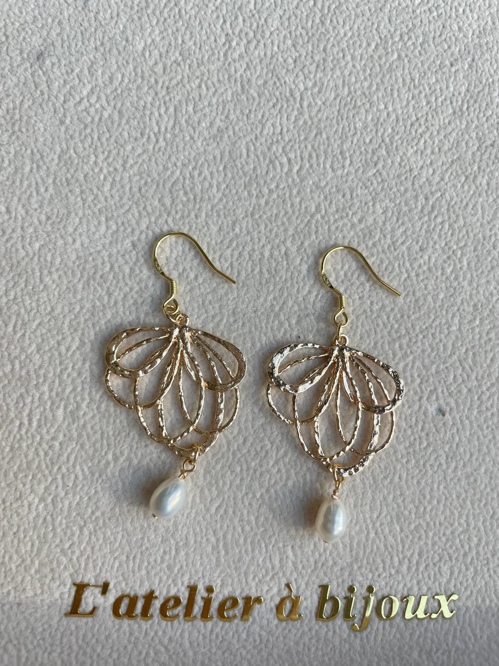 A pair of gold-tone dangling earrings with an openwork leaf or fan pattern and a small white pearl drop, displayed on a textured off-white surface with the text 'L'atelier à bijoux' at the bottom.