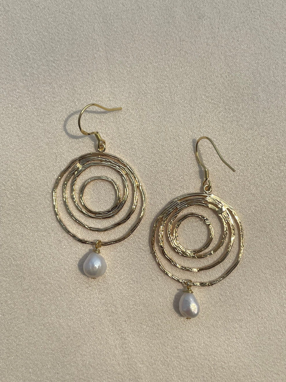 A pair of gold-toned spiral drop earrings with a small white teardrop pearl hanging from each spiral, displayed on a textured beige background.