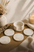 A set of round off-white marble coasters and a matching marble cup or candle holder arranged on a wooden surface with a draped cream cloth, dried flowers in a vase, and a lit candle in the background.
