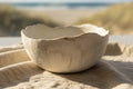 A handmade ceramic bowl with a scalloped rim and a textured, matte off-white finish, resting on a beige linen cloth with a blurred beach background.