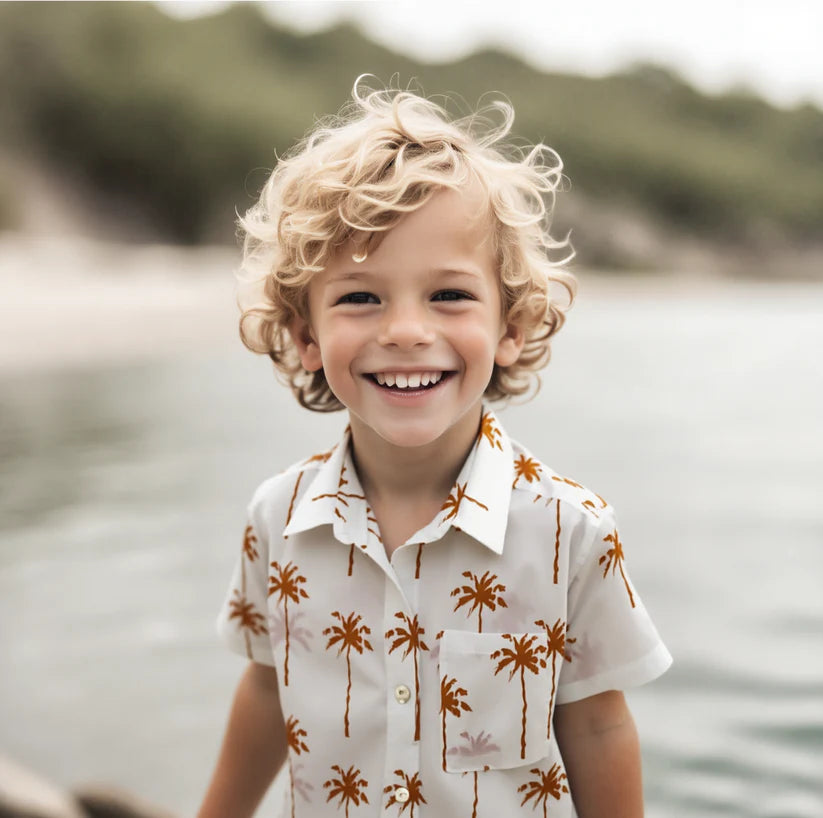 A young child with curly blond hair smiling outdoors by the water, wearing a white short-sleeve button-up shirt with a brown palm tree print.