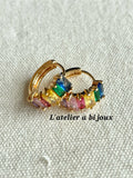 A pair of gold hoop earrings with colorful rectangular gemstones in yellow, green, blue, pink, and purple, displayed on a textured off-white fabric background with the text 'L'atelier à bijoux'.