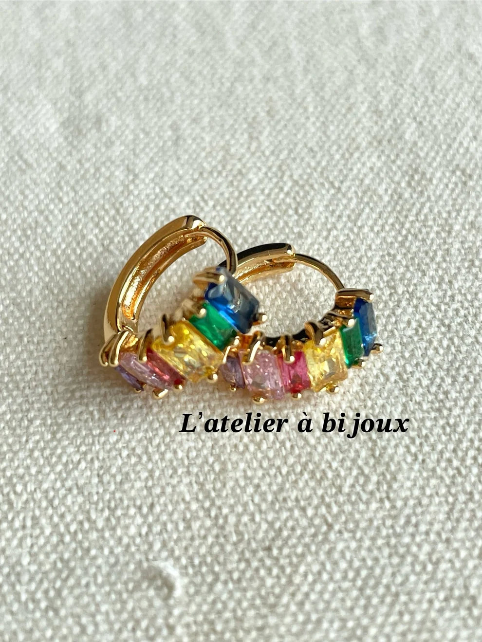 A pair of gold hoop earrings with colorful rectangular gemstones in yellow, green, blue, pink, and purple, displayed on a textured off-white fabric background with the text 'L'atelier à bijoux'.