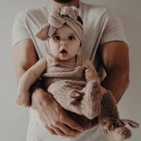 A baby wearing a knitted beige outfit and matching headband is held in the arms of an adult wearing a white t-shirt against a plain light background.