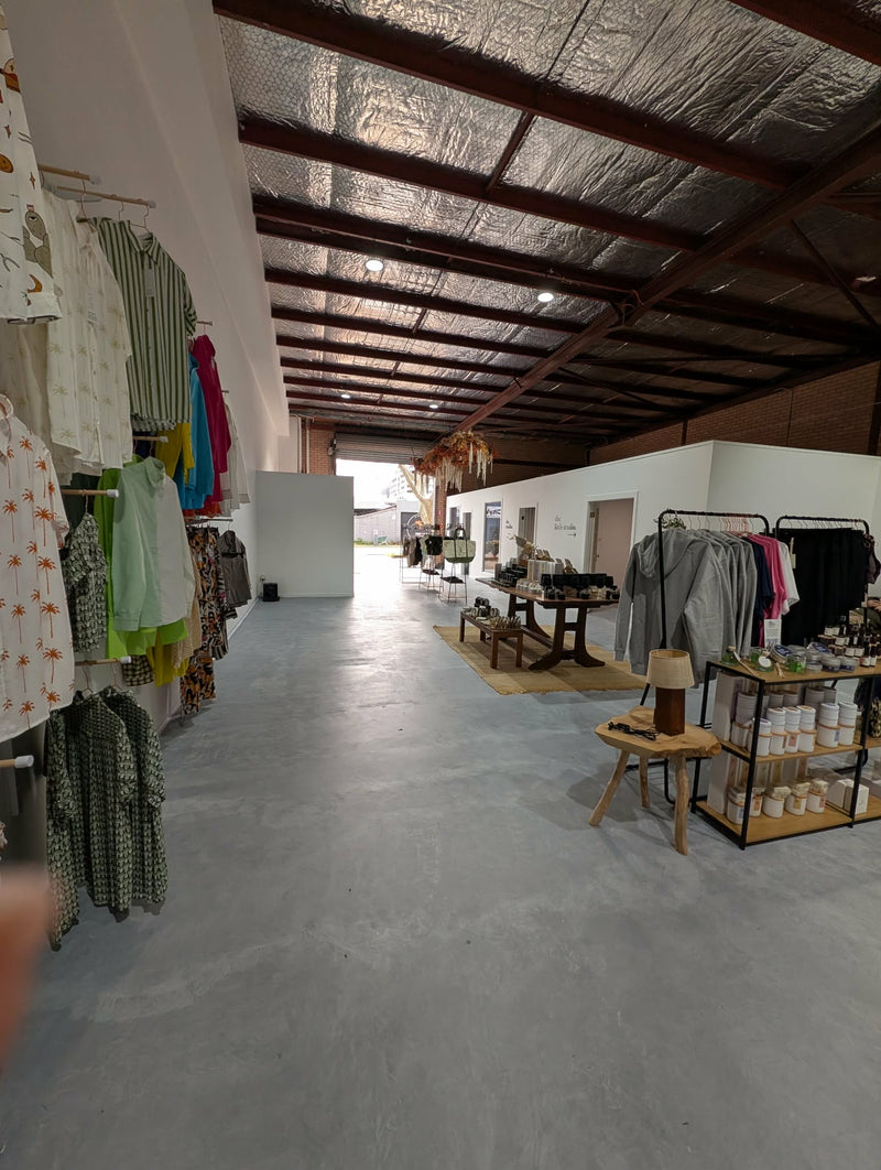 A wide interior view of a clothing boutique or retail space with a concrete floor, exposed ceiling beams, and racks of garments on both sides. Shelves and tables with merchandise are visible on the right side.