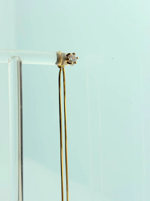A gold chain necklace with a small gold ring setting holding a clear gemstone, displayed on a clear acrylic stand against a pale blue background.