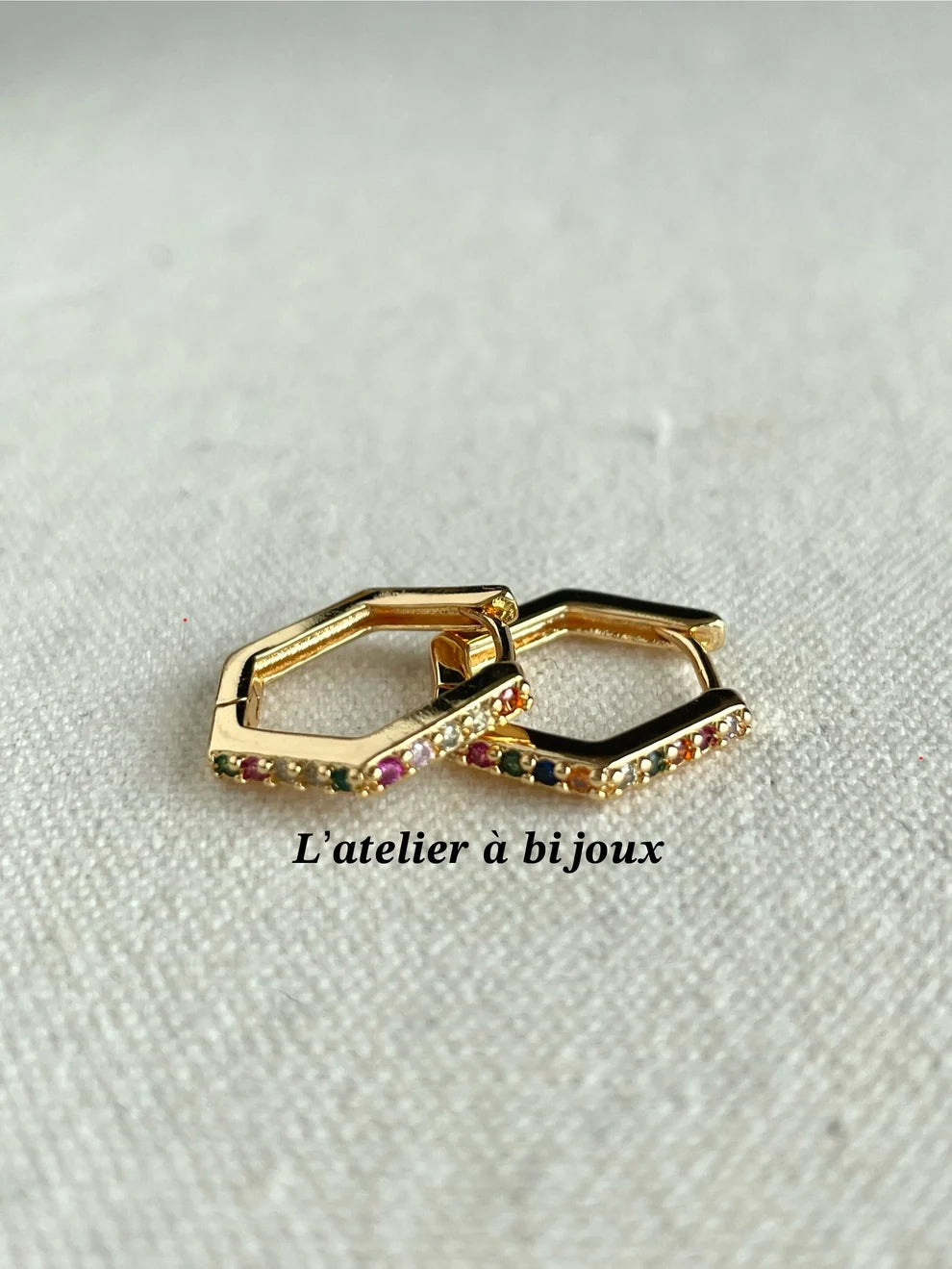 A pair of gold-toned geometric hoop earrings with small multicolor gemstones, displayed on a textured off-white fabric background with the text 'L'atelier à bijoux' printed below.