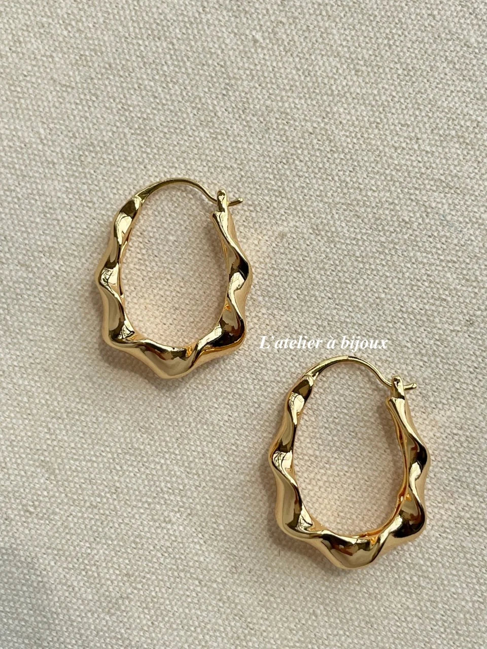 A pair of gold-toned, organic-shaped hoop earrings with a wavy, textured finish, displayed on a neutral beige fabric background.