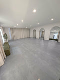 A large, empty studio room with a gray concrete floor, white walls, recessed ceiling lights, cream curtains, and arched mirrors. A small green bench and potted plant are visible on the left side.