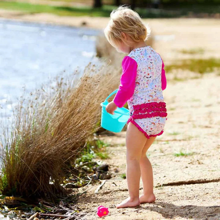 A toddler standing on a sandy lakeshore wearing a floral long-sleeve swim shirt with bright pink sleeves and matching swim trunks, holding a small blue bucket.