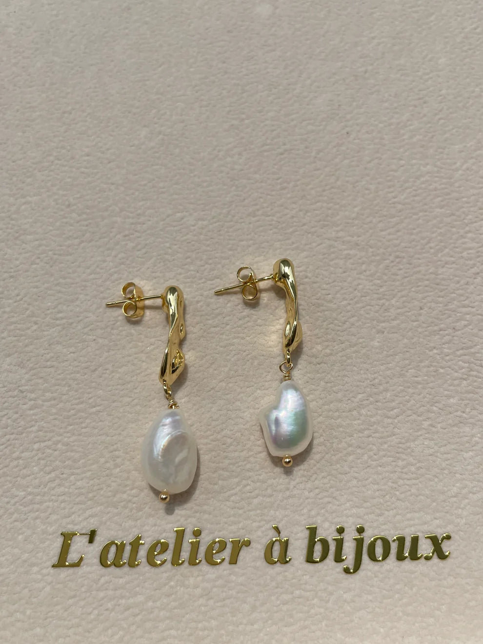 A pair of gold huggie hoop earrings with dangling baroque pearl drops, displayed on a textured beige surface with the French text 'L'atelier à bijoux' printed below.