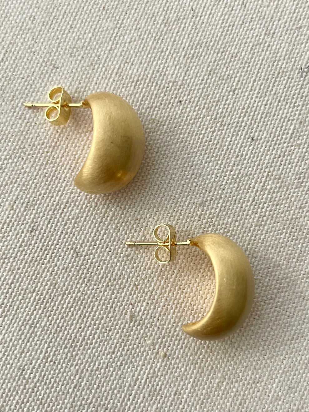 A pair of gold crescent moon stud earrings with butterfly backings, displayed on a textured beige fabric background.