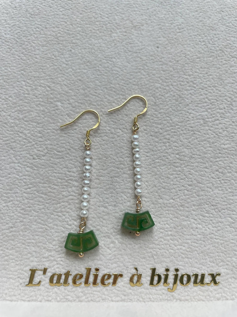 A pair of gold-tone drop earrings with small white pearl-like beads and green fan-shaped charms, displayed on a textured light background with the text 'L'atelier à bijoux' at the bottom.