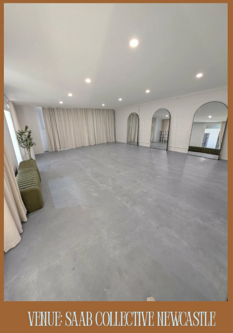A large, empty studio or dance room with a smooth gray concrete floor, white walls, arched mirrors, and beige curtains. A green upholstered bench sits near the left window, and the photo is framed by a brown border with the text 'VENUE-SAAB COLLECTIVE NEWCASTLE' at the bottom.
