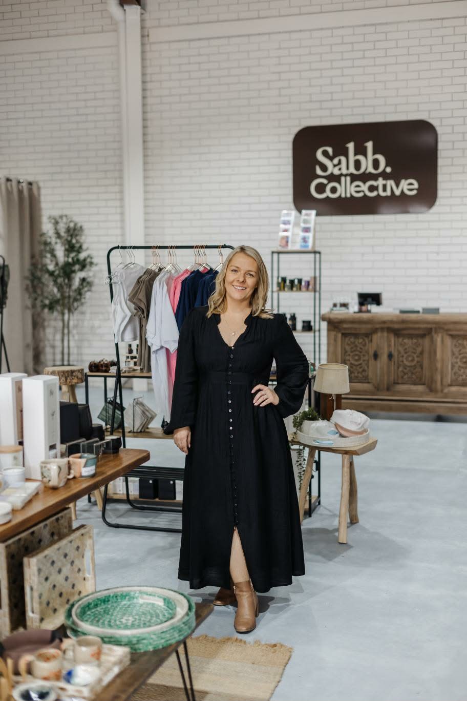 A woman stands in a boutique interior wearing a long black button-front dress. The space has white brick walls, a wooden sign reading 'Sabb Collective,' clothing racks, tables, and decor.
