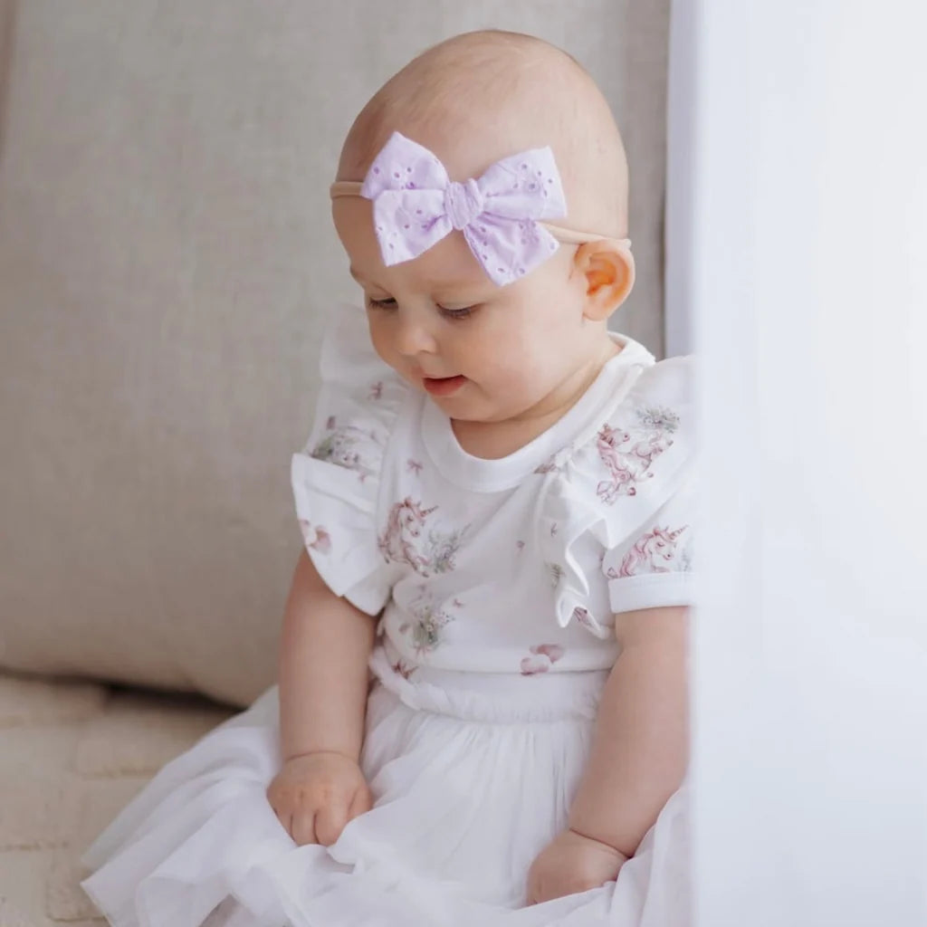 A baby girl wearing a white short-sleeve dress with a delicate pink floral print and a matching white ruffled bow headband with a light purple polka-dot bow, sitting in front of a soft beige cushion.