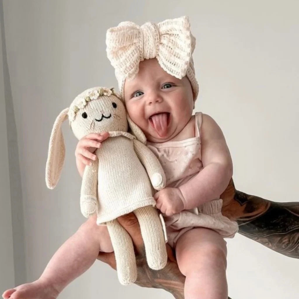 A baby wearing a cream knit bow headband and matching cream outfit, holding a cream knitted bunny doll while sitting on an adult's lap against a soft light background.