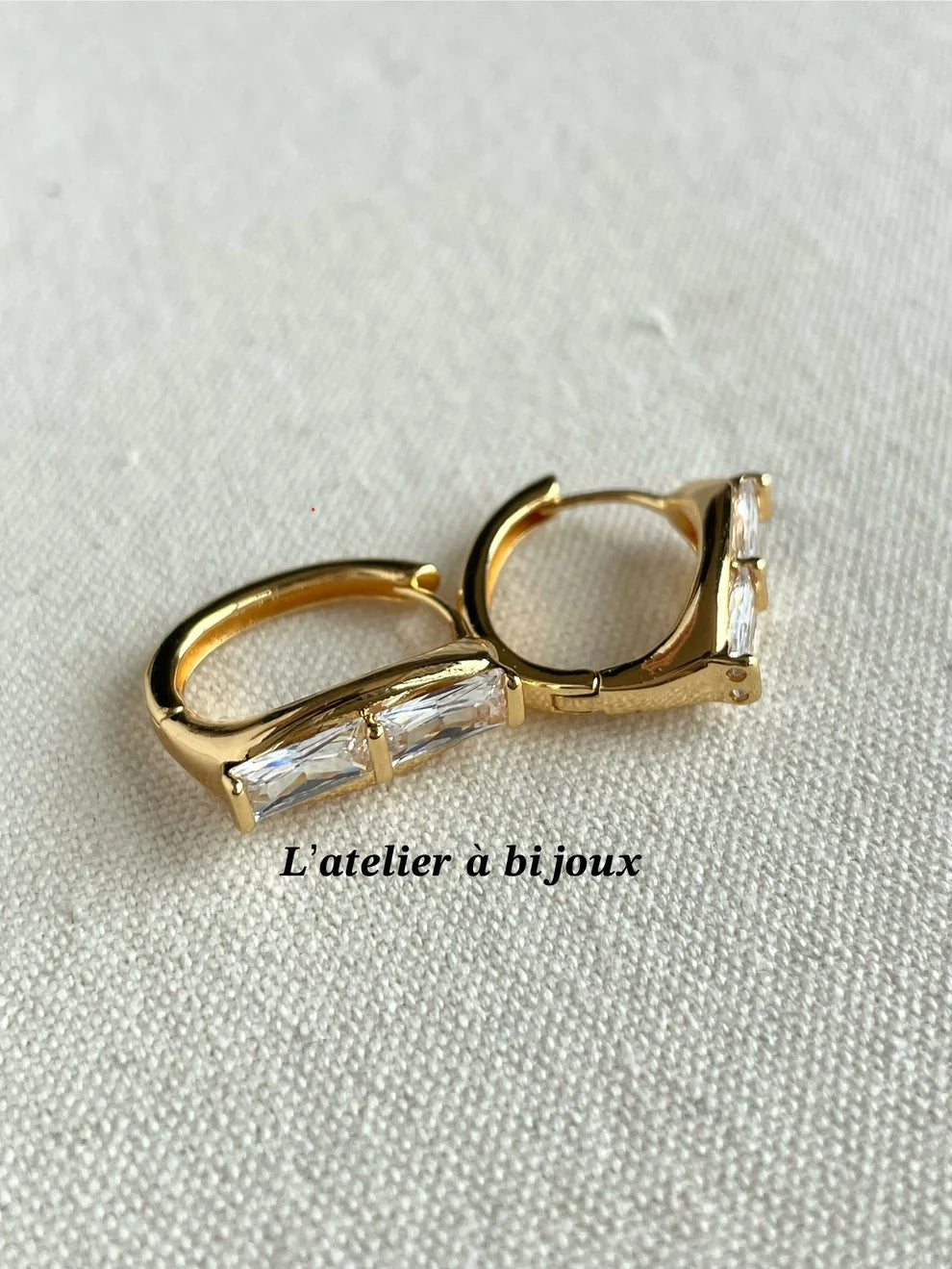 A pair of gold huggie hoop earrings with baguette-cut clear crystals, displayed on a textured off-white fabric background with a watermark overlay.