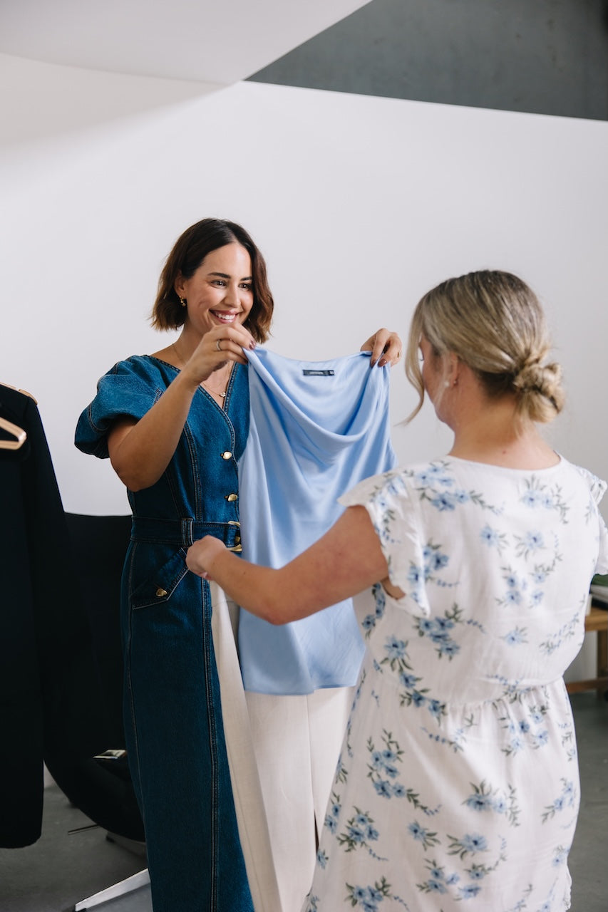 A woman in a blue denim dress holds up a light blue top for another woman wearing a white floral dress, in a minimalist studio setting.