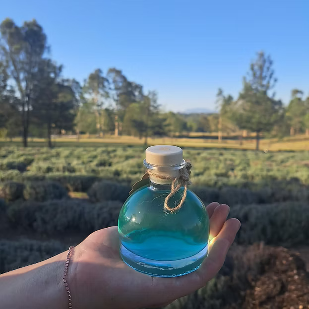 A hand holding a small glass bottle filled with bright blue liquid, set against a blurred outdoor background of trees and fields under a clear blue sky.