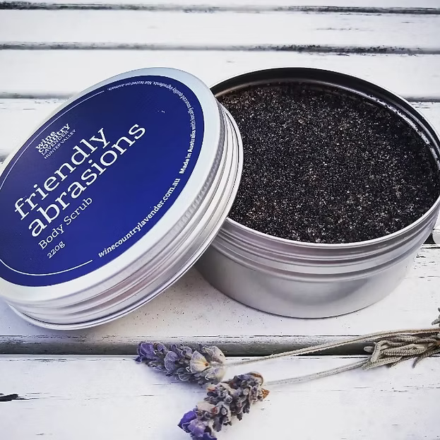 An open round silver tin of body scrub with the lid leaning beside it, showing a dark charcoal scrub inside. The lid label reads 'friendly abrasions' and 'Body scrub 220g'.