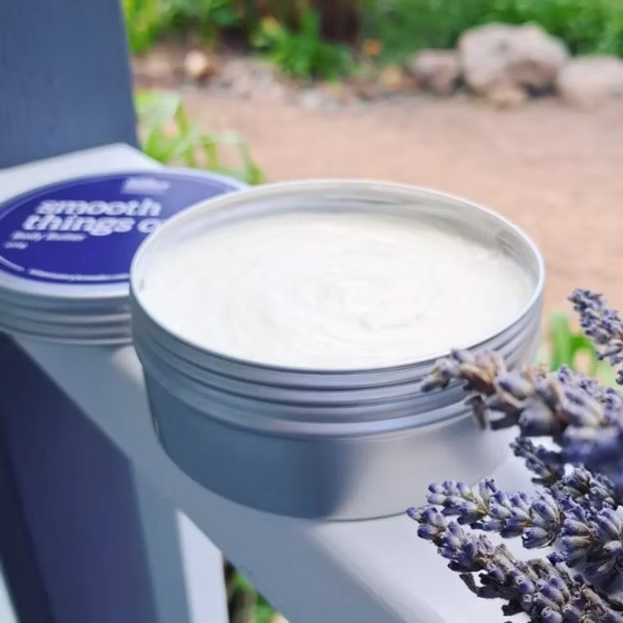 An open round metal tin of body butter showing the white cream inside, with a matching stacked tin in the background and lavender sprigs in the foreground on a white surface.