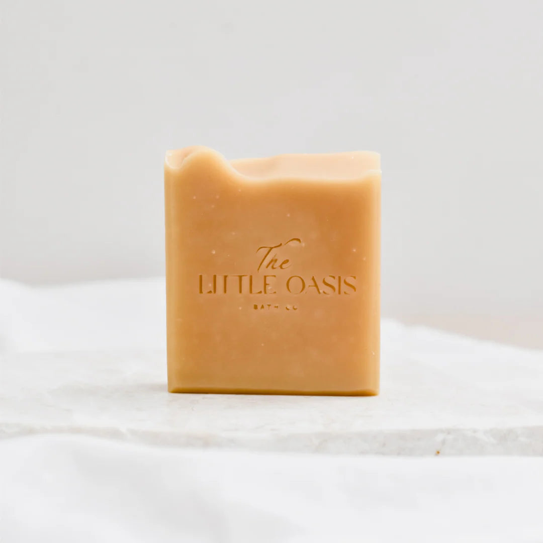 A rectangular bar of warm tan soap with a wavy top edge, embossed with the brand name 'The LITTLE OASIS BATH CO'. The soap is displayed on a white marble slab against a soft, light gray background.