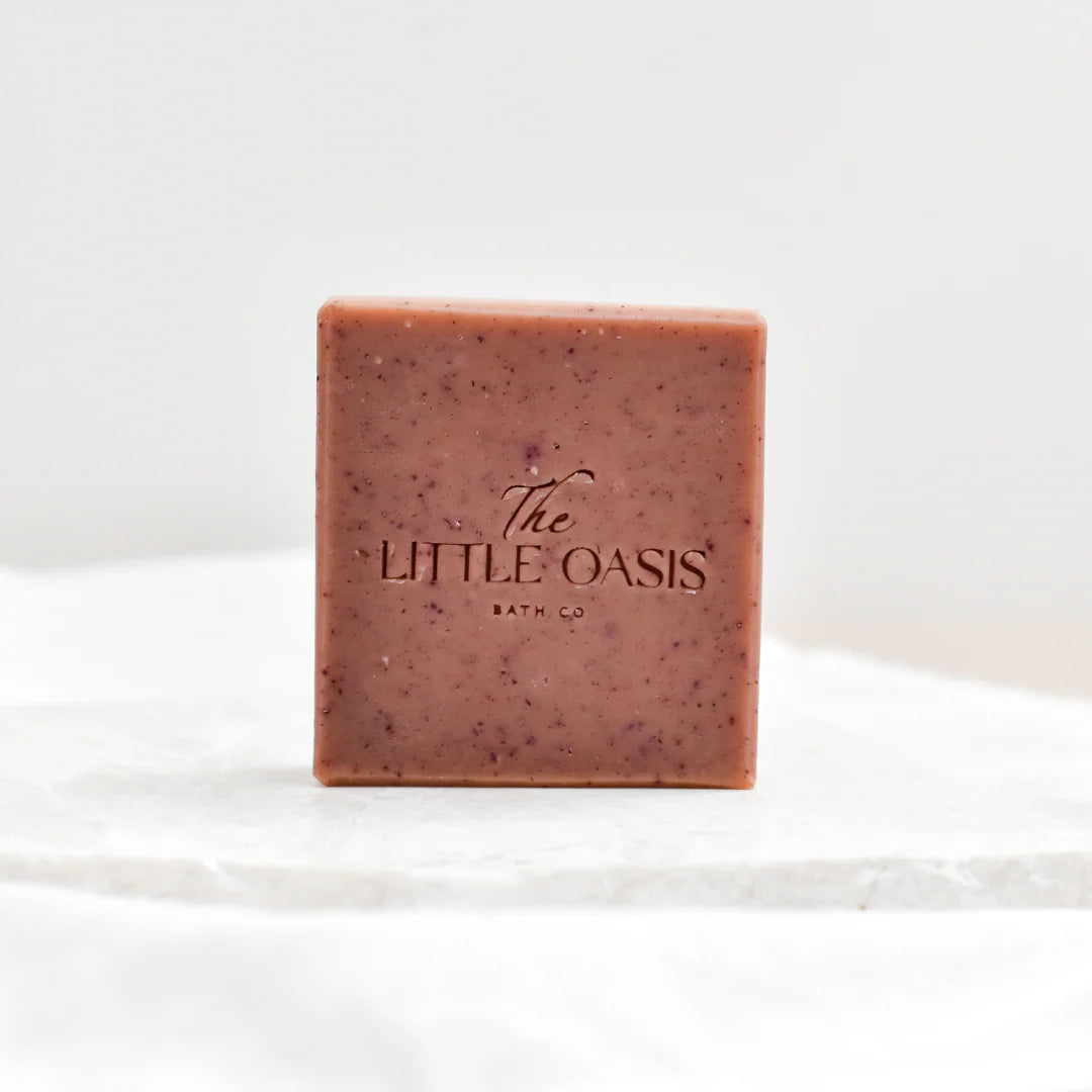 A square bar of bath soap from The Little Oasis Bath Co, featuring a dusty rose pink color with dark purple speckles, standing upright on a white marble slab against a white background.